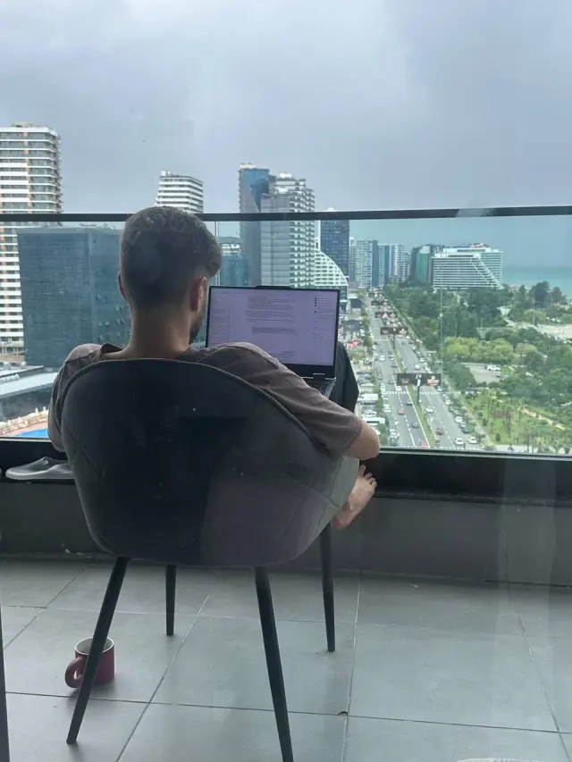 Alexey working on a laptop by a balcony overlooking a city